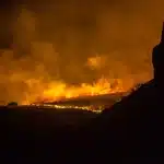 Firefighters spraying water on a large wildfire at night with orange flames and thick smoke