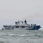 Aircraft carrier approaching a tropical port with sailors visible on deck